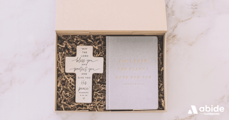 A cross with a blessing on it next to a cloth journal on a bed of packing.