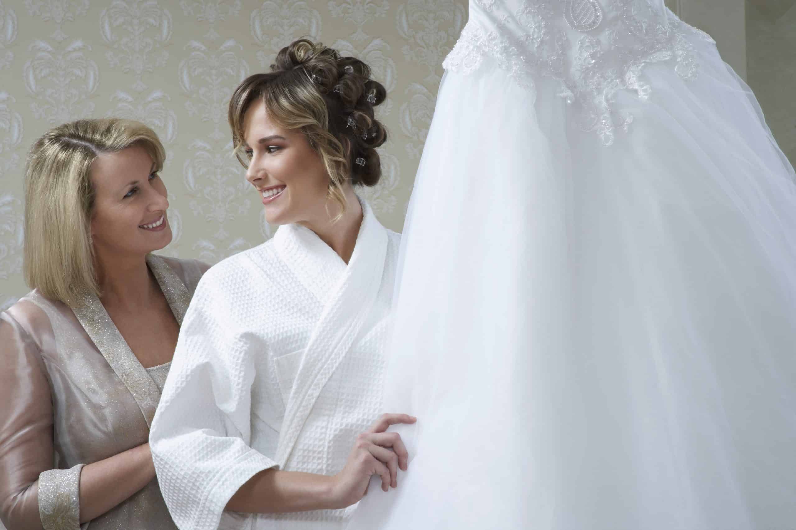 Bride in bathrobe touching wedding dress mother looking on